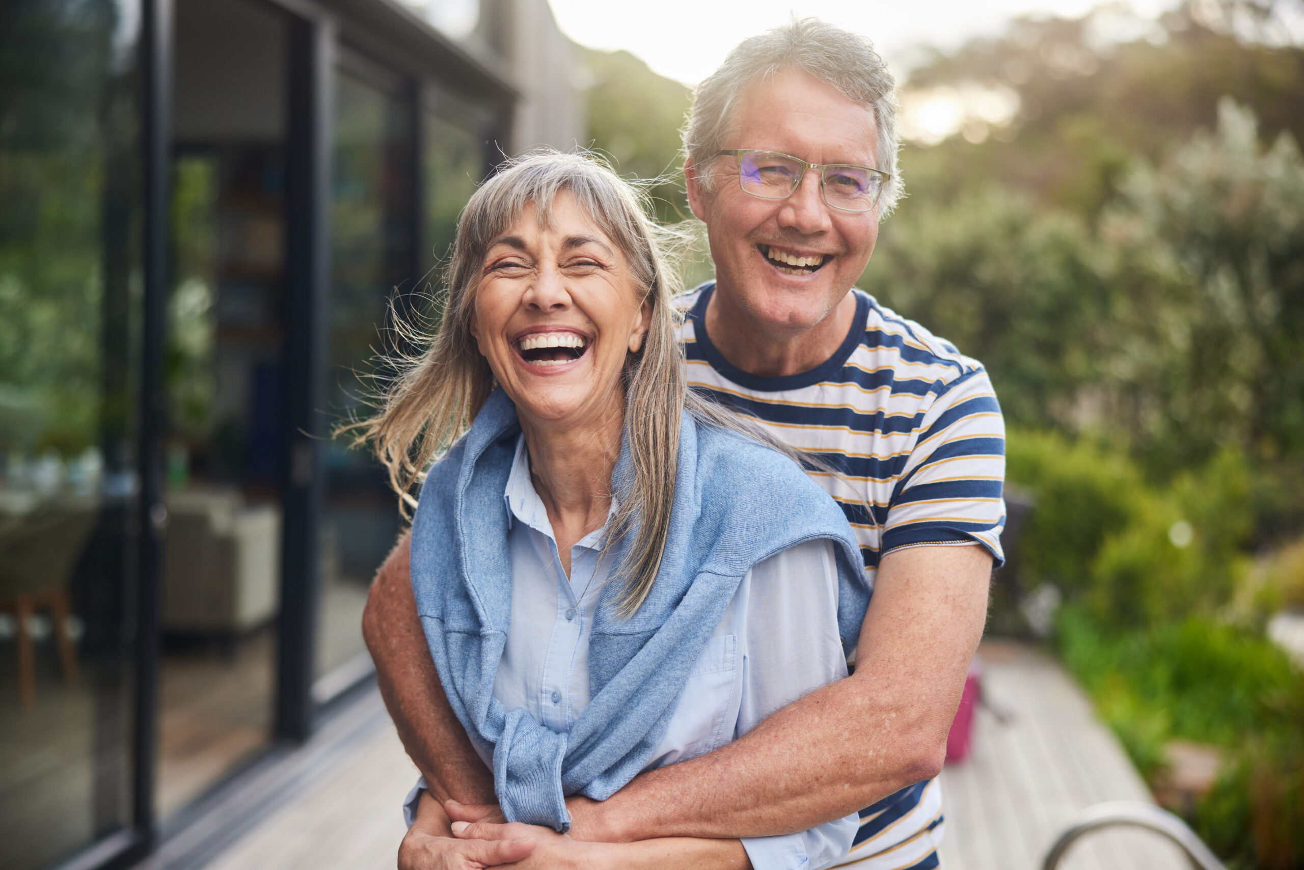 Mature couple standing arm in arm together and laughing outside in the back yard at home in summer Mature Couple Standing Arm In Arm And Laughing Outside In Summer